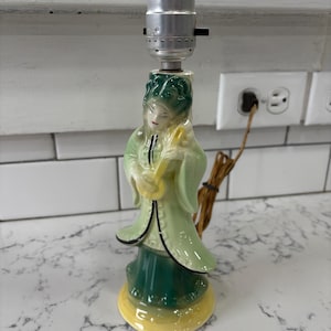 May include: A ceramic lamp base featuring a woman in a green and yellow robe playing a musical instrument. The lamp base is in the style of a traditional Asian figurine.