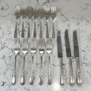 May include: A collection of antique silverware, including forks and knives, arranged on a marble countertop. The silverware features ornate floral detailing on the handles and tines. The knives have a smooth, polished blade.