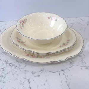 May include: A cream-colored porcelain dish set with a floral pattern. The set includes a large plate, a smaller plate, and a bowl. Each piece has a scalloped edge and a silver trim. The floral design features pink flowers.