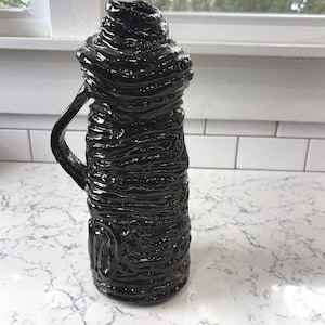 May include: A glossy black ceramic pitcher with a textured, layered design. The pitcher has a handle and a wide, irregular opening. The surface reflects light, highlighting the unique, handcrafted appearance. The pitcher is on a white countertop.