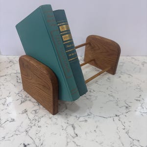 May include: A wooden book holder with two teal-colored books. The book holder is made of light brown wood and has a simple, minimalist design. The books have gold lettering and are titled "The Heritage" and "Silver Nutmeg".