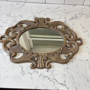 May include: Ornate, oval-shaped mirror with a bronze-colored frame. The frame features detailed scrollwork and a central oval mirror. The mirror is a decorative piece, suitable for wall decor or vanity use.