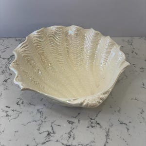 May include: A large, pearlescent white ceramic seashell-shaped bowl. The bowl has a wavy, textured interior and a scalloped edge. The bowl is designed to resemble a clam shell and is suitable for decorative purposes.