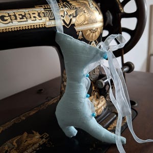 May include: A light blue fabric shoe with blue beads and a white ribbon hanging from an antique sewing machine.