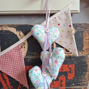 May include: Three heart-shaped fabric decorations with floral patterns in blue, pink, and yellow, hanging by lavender ribbons. Each heart has a decorative button. The background features a wooden surface and bunting.
