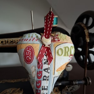 May include: A heart-shaped fabric cushion with a red ribbon and a small white chicken charm. The cushion is decorated with a vintage-style print featuring the words "Free Range Eggs" and "Orchard".