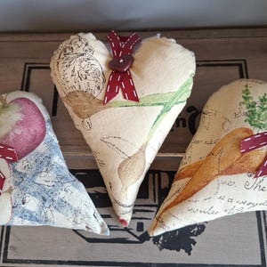 May include: Three heart-shaped fabric ornaments with printed designs and burgundy ribbon accents. One features an apple, another a bird, and the third carrots. Each has a burgundy ribbon and button for hanging. The ornaments are on a wooden surface.