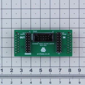 May include: A green circuit board with black connectors and the text "EXCHANGE TURING MACHINE ADAPTER REV2 2025". The board has "OUT" and "IN" labels, and the website archaea.co.uk is printed on the board.