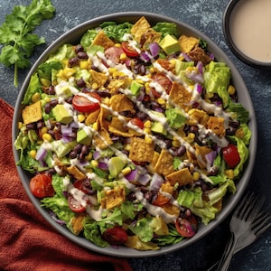 May include: A large salad in a gray bowl, featuring romaine lettuce, black beans, corn, avocado, red onion, and cherry tomatoes. Tortilla chips are scattered throughout, with a creamy dressing drizzled over the top. A side of dressing is in a black bowl.