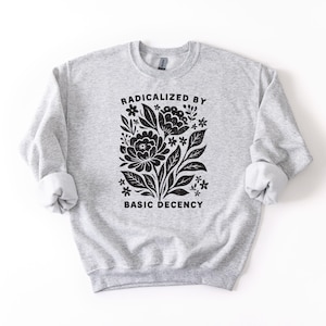 May include: A light gray sweatshirt with rolled-up sleeves. The front features a black floral design with the words "RADICALIZED BY BASIC DECENCY" in a bold, sans-serif font. The sweatshirt is made of a soft, comfortable material.