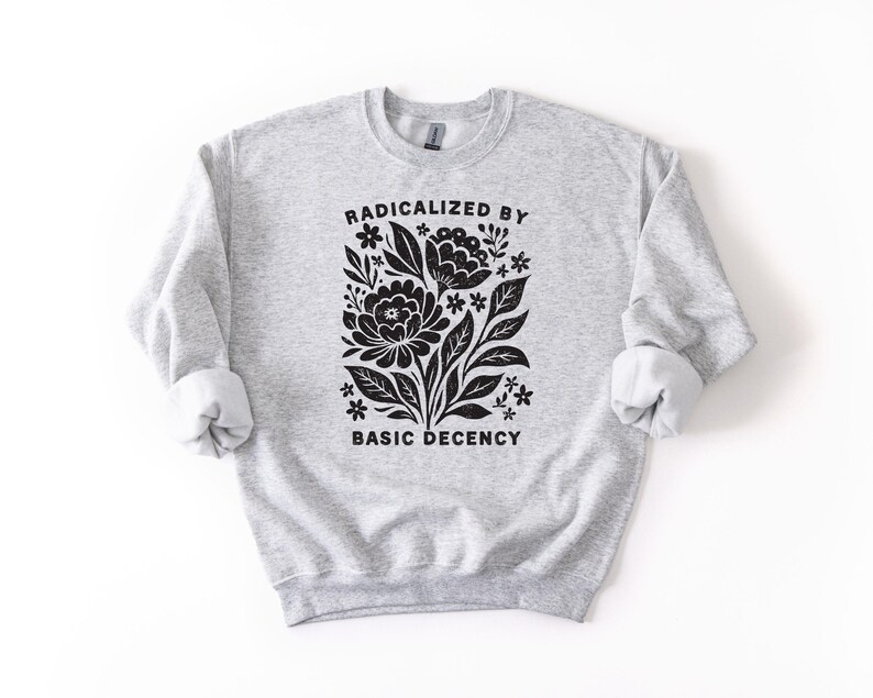 May include: A light gray sweatshirt with rolled-up sleeves. The front features a black floral design with the words "RADICALIZED BY BASIC DECENCY" in a bold, sans-serif font. The sweatshirt is made of a soft, comfortable material.