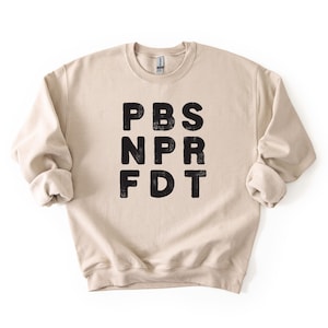 May include: A beige crewneck sweatshirt with the letters PBS, NPR, and FDT printed in black, stacked vertically. The sweatshirt has long sleeves with rolled cuffs and a ribbed neckline.