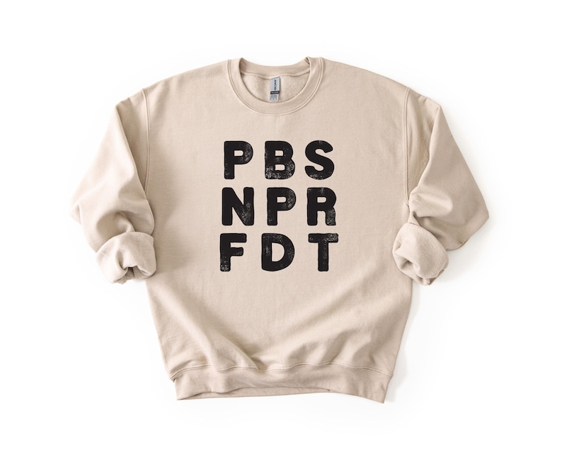 May include: A beige crewneck sweatshirt with the letters PBS, NPR, and FDT printed in black, stacked vertically. The sweatshirt has long sleeves with rolled cuffs and a ribbed neckline.
