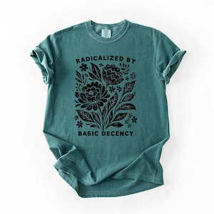 May include: Teal-colored t-shirt with a black floral design and the text "RADICALIZED BY BASIC DECENCY." The graphic features stylized flowers, leaves, and other botanical elements. The shirt has short sleeves and a crew neck.