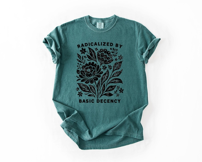 May include: Teal-colored t-shirt with a black floral design and the text "RADICALIZED BY BASIC DECENCY." The graphic features stylized flowers, leaves, and other botanical elements. The shirt has short sleeves and a crew neck.