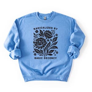 May include: A light blue sweatshirt with the text "RADICALIZED BY BASIC DECENCY" in black, along with a floral design. The design features stylized flowers and leaves. The sweatshirt has long sleeves and a crew neck.