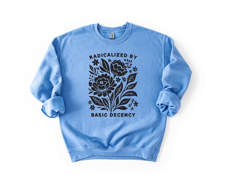 May include: A light blue sweatshirt with the text "RADICALIZED BY BASIC DECENCY" in black, along with a floral design. The design features stylized flowers and leaves. The sweatshirt has long sleeves and a crew neck.