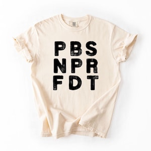 May include: Cream-colored t-shirt with the black text "PBS NPR FDT" printed on the front. The shirt has short sleeves and a crew neck. The fabric appears to be soft and comfortable.