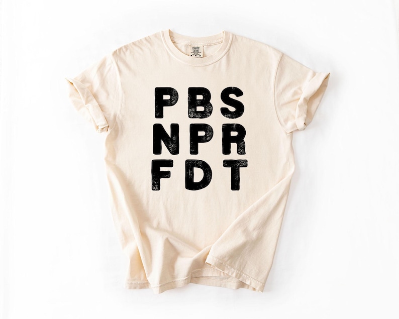 May include: Cream-colored t-shirt with the black text "PBS NPR FDT" printed on the front. The shirt has short sleeves and a crew neck. The fabric appears to be soft and comfortable.