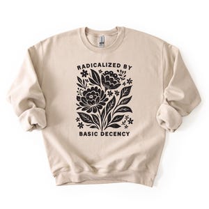 May include: A beige crewneck sweatshirt with the words "RADICALIZED BY BASIC DECENCY" printed in black. The design features a floral graphic in black, including flowers, leaves, and stems.