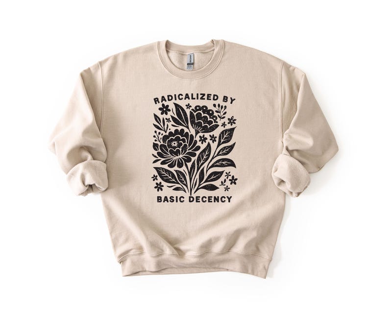 May include: A beige crewneck sweatshirt with the words "RADICALIZED BY BASIC DECENCY" printed in black. The design features a floral graphic in black, including flowers, leaves, and stems.