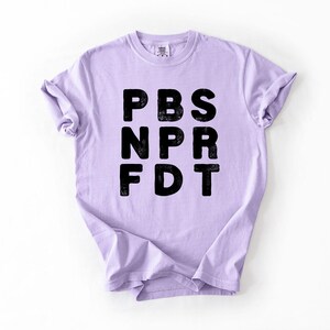 May include: A light purple t-shirt with the letters PBS, NPR, and FDT printed in black. The letters have a distressed, textured appearance. The shirt is made of a soft material and has rolled-up sleeves.