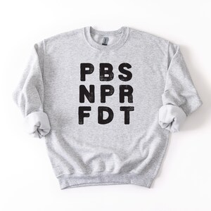 May include: A light gray sweatshirt with the letters PBS, NPR, and FDT printed in black. The sweatshirt has a crew neck and long sleeves with rolled-up cuffs. The garment is made of a soft, comfortable material.