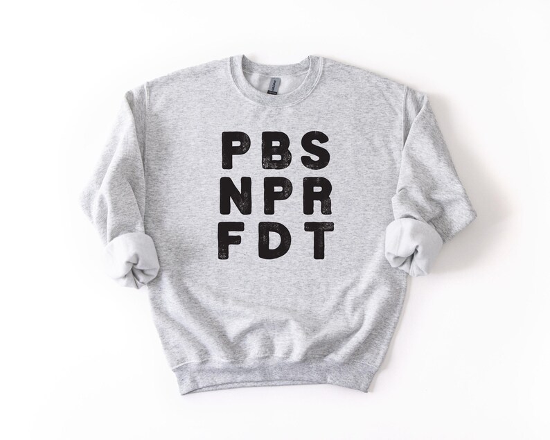 May include: A light gray sweatshirt with the letters PBS, NPR, and FDT printed in black. The sweatshirt has a crew neck and long sleeves with rolled-up cuffs. The garment is made of a soft, comfortable material.