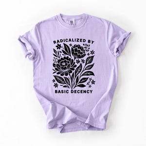 May include: A light purple t-shirt with the text "RADICALIZED BY BASIC DECENCY" in black. The shirt features a black floral design with leaves and flowers. The t-shirt is made of a soft material.