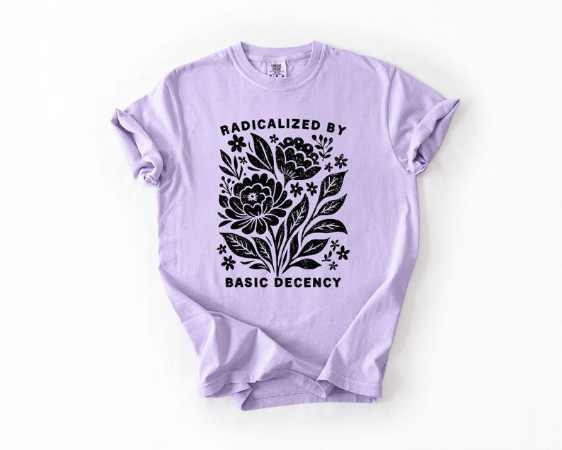 May include: A light purple t-shirt with the text "RADICALIZED BY BASIC DECENCY" in black. The shirt features a black floral design with leaves and flowers. The t-shirt is made of a soft material.