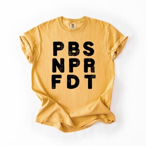 May include: Mustard yellow t-shirt with the black text "PBS NPR FDT" printed on the front. The shirt has short sleeves and a crew neck. The fabric appears to be soft and comfortable, with a slightly faded look.