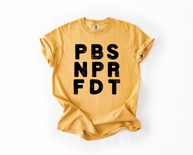 May include: Mustard yellow t-shirt with the black text "PBS NPR FDT" printed on the front. The shirt has short sleeves and a crew neck. The fabric appears to be soft and comfortable, with a slightly faded look.