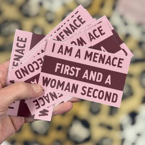 May include: A stack of pink rectangular stickers with the text "I AM A MENACE FIRST AND A WOMAN SECOND" in burgundy. The word "MENACE" is also printed on the top and bottom of the stickers. The background is a blurred leopard print.