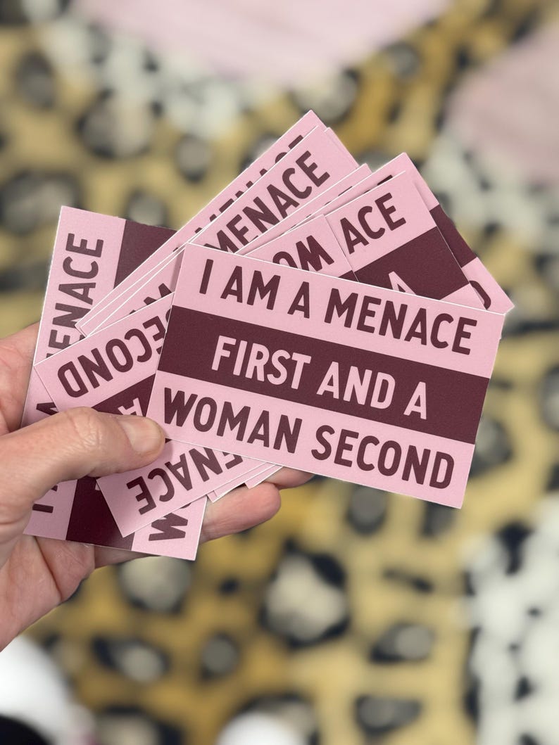 May include: A stack of pink rectangular stickers with the text "I AM A MENACE FIRST AND A WOMAN SECOND" in burgundy. The word "MENACE" is also printed on the top and bottom of the stickers. The background is a blurred leopard print.