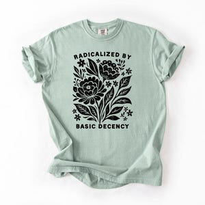 May include: A light green t-shirt with rolled-up sleeves. The shirt features a black floral design with the words "RADICALIZED BY BASIC DECENCY" in a black, bold font.