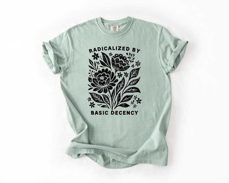 May include: A light green t-shirt with rolled-up sleeves. The shirt features a black floral design with the words "RADICALIZED BY BASIC DECENCY" in a black, bold font.