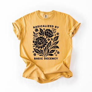May include: Mustard yellow t-shirt with rolled sleeves. The front features a black floral design with the words "RADICALIZED BY BASIC DECENCY" in a bold, sans-serif font. The shirt is made of a soft, comfortable material.