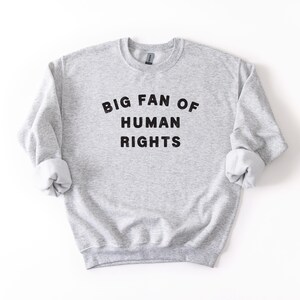 May include: A light gray sweatshirt with the text "BIG FAN OF HUMAN RIGHTS" in black block letters. The sweatshirt has a crew neck and long sleeves with rolled-up cuffs. The garment is made of a soft, comfortable material.