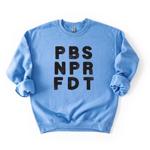 May include: A light blue sweatshirt with the letters PBS, NPR, and FDT printed in black. The sweatshirt has long sleeves and a crew neck. The image is a close-up shot of the sweatshirt against a white background.