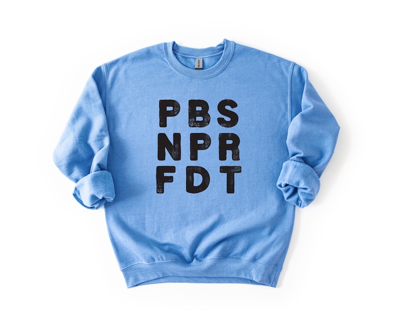 May include: A light blue sweatshirt with the letters PBS, NPR, and FDT printed in black. The sweatshirt has long sleeves and a crew neck. The image is a close-up shot of the sweatshirt against a white background.