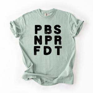 May include: A light green t-shirt with the black letters PBS, NPR, and FDT printed on the front. The shirt has short sleeves and a crew neck. The fabric appears to be soft and comfortable.