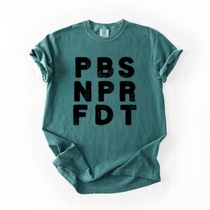 May include: Teal-colored t-shirt with the black text "PBS NPR FDT" printed vertically. The shirt has short sleeves and a crew neck. The fabric appears soft and slightly faded.