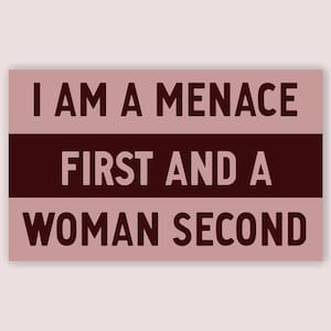 May include: A rectangular sign with the text "I AM A MENACE FIRST AND A WOMAN SECOND" in dark brown letters. The sign has a light pink background with a dark brown horizontal stripe in the middle.