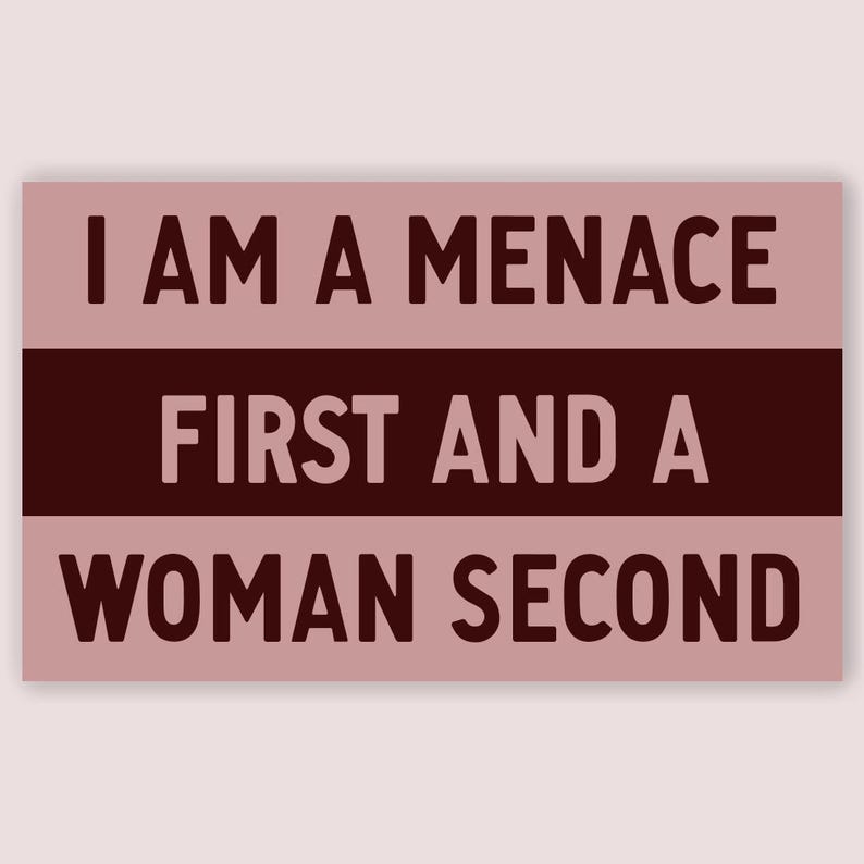 May include: A rectangular sign with the text "I AM A MENACE FIRST AND A WOMAN SECOND" in dark brown letters. The sign has a light pink background with a dark brown horizontal stripe in the middle.