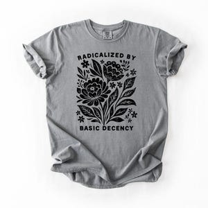 May include: A gray t-shirt with a black floral design and the text "RADICALIZED BY BASIC DECENCY". The graphic features stylized flowers, leaves, and small star-like shapes. The shirt has rolled-up sleeves and a crew neck.