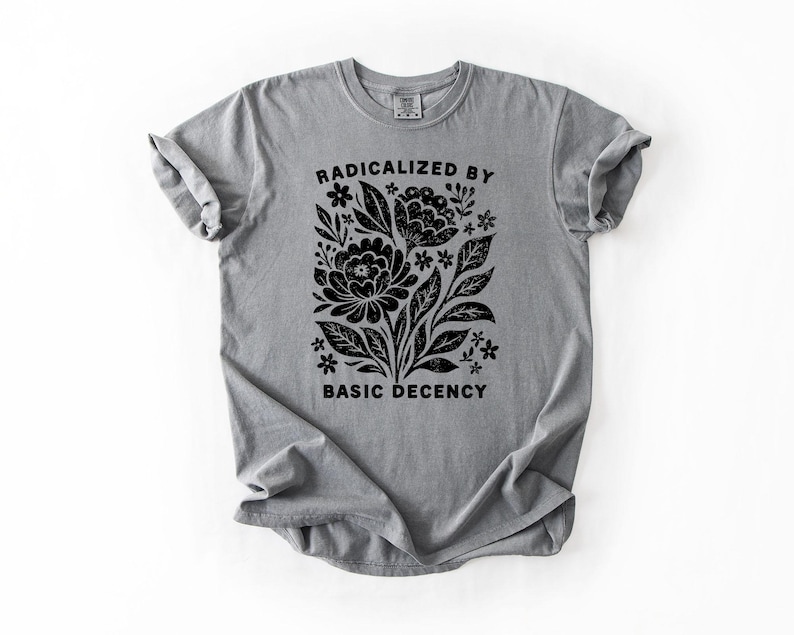 May include: A gray t-shirt with a black floral design and the text "RADICALIZED BY BASIC DECENCY". The graphic features stylized flowers, leaves, and small star-like shapes. The shirt has rolled-up sleeves and a crew neck.