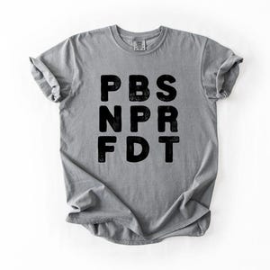 May include: A gray t-shirt with the black text "PBS NPR FDT" printed on the front. The shirt is made of a soft material and has rolled-up sleeves. The shirt is displayed on a white background.