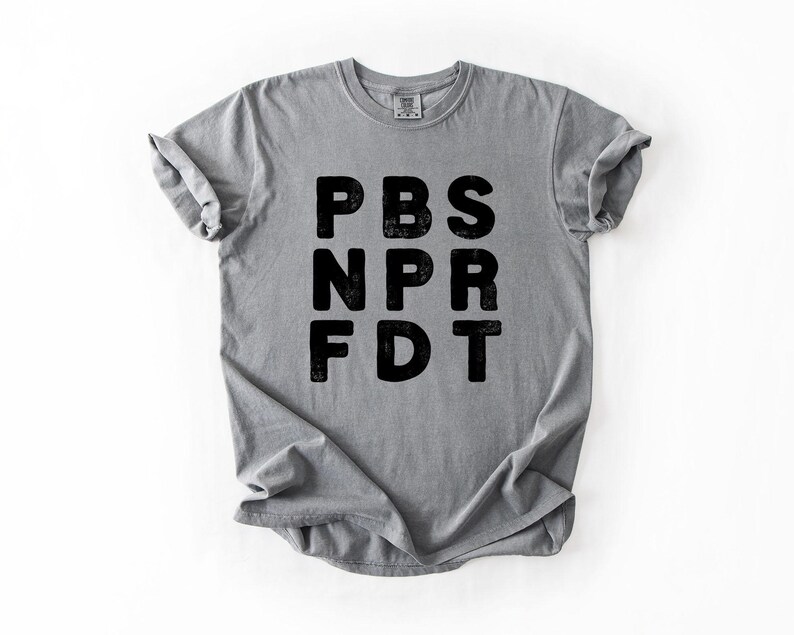 May include: A gray t-shirt with the black text "PBS NPR FDT" printed on the front. The shirt is made of a soft material and has rolled-up sleeves. The shirt is displayed on a white background.