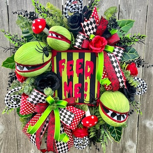 May include: A decorative wreath featuring a central sign with the text FEED ME! in red, surrounded by green and red monster plant heads, black and white checkered ribbons, red polka dot ribbons, red and black roses, and small red mushrooms.