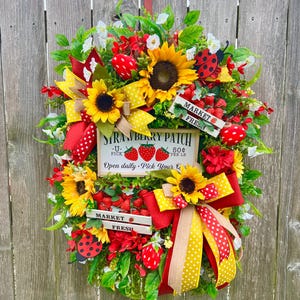 May include: A vibrant wreath featuring sunflowers, red berries, and green foliage. The wreath includes a sign that reads "Strawberry Patch" and "Market Fresh". Red and yellow ribbons and ladybug accents add to the decorative design.